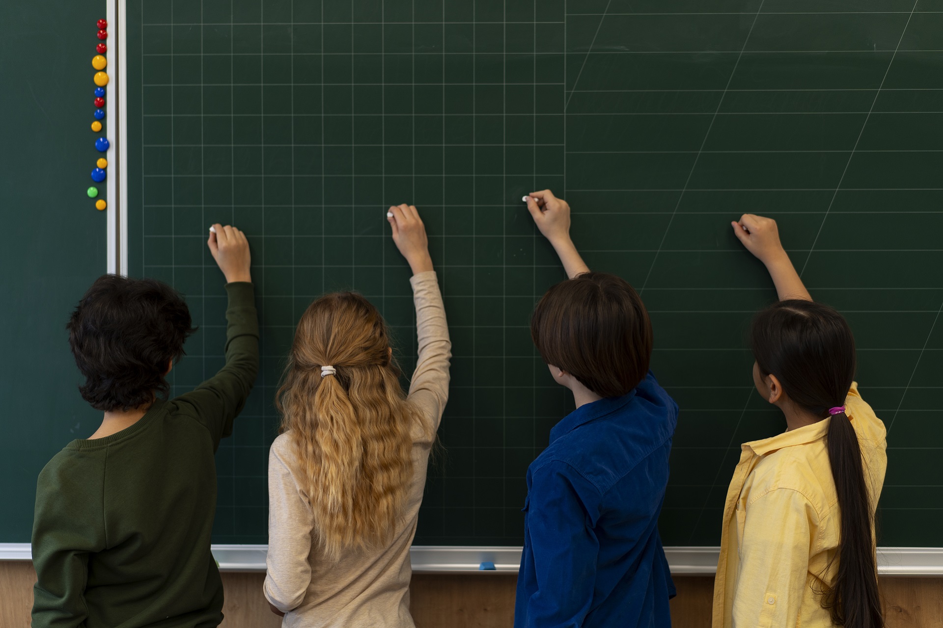 quatro crianças de costas para a foto, escrevem no quadro negro da sala de aula. Duas meninas e dois meninos