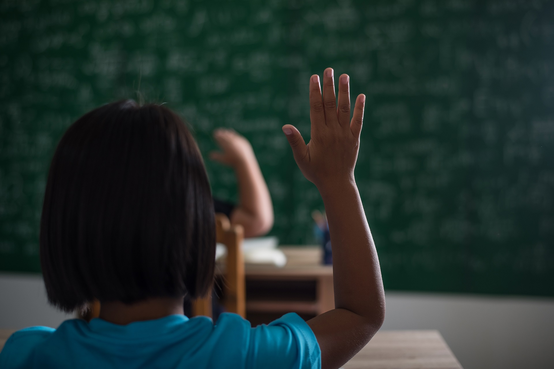 Criança de pele escura, sentada numa cadeira, de costas, levantando a mão dentro da sala de aula, de frente para o quadro