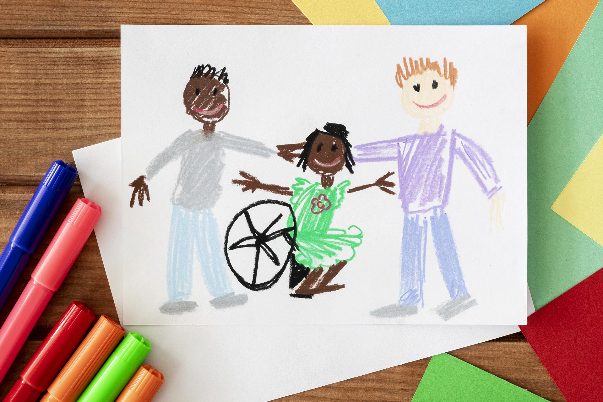 desenho feito a mão com canetas coloridas, onde está ilustrada uma família composta por dois pais de etnias diferentes e uma criança negra sentada em uma cadeira de rodas. Ambos sorriem e se abraçam